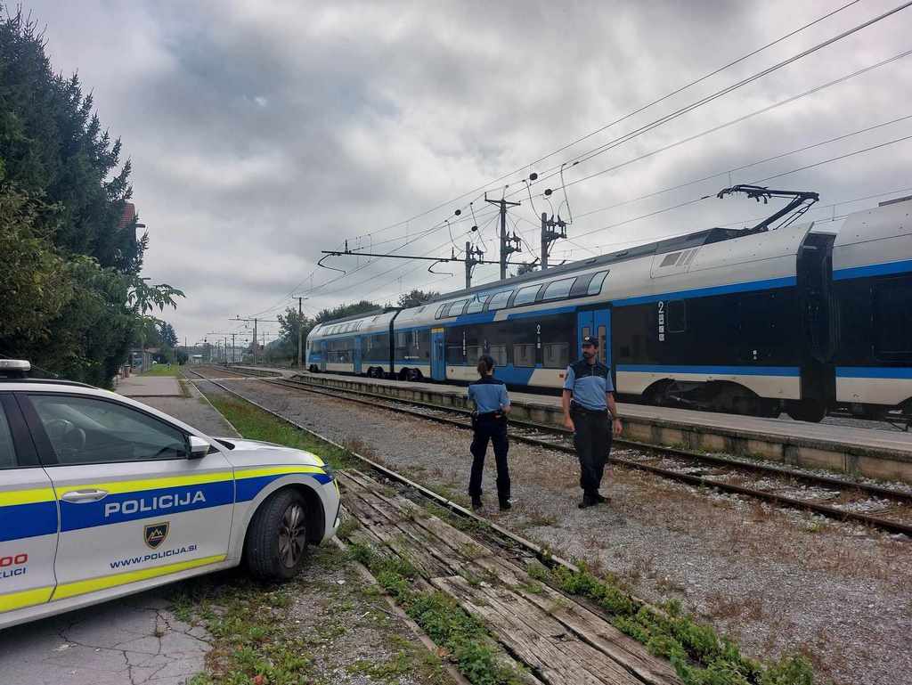 Policista in policijsko vozilo stojijo ob železniški progi, po kateri se ravno mimo pelje vlak