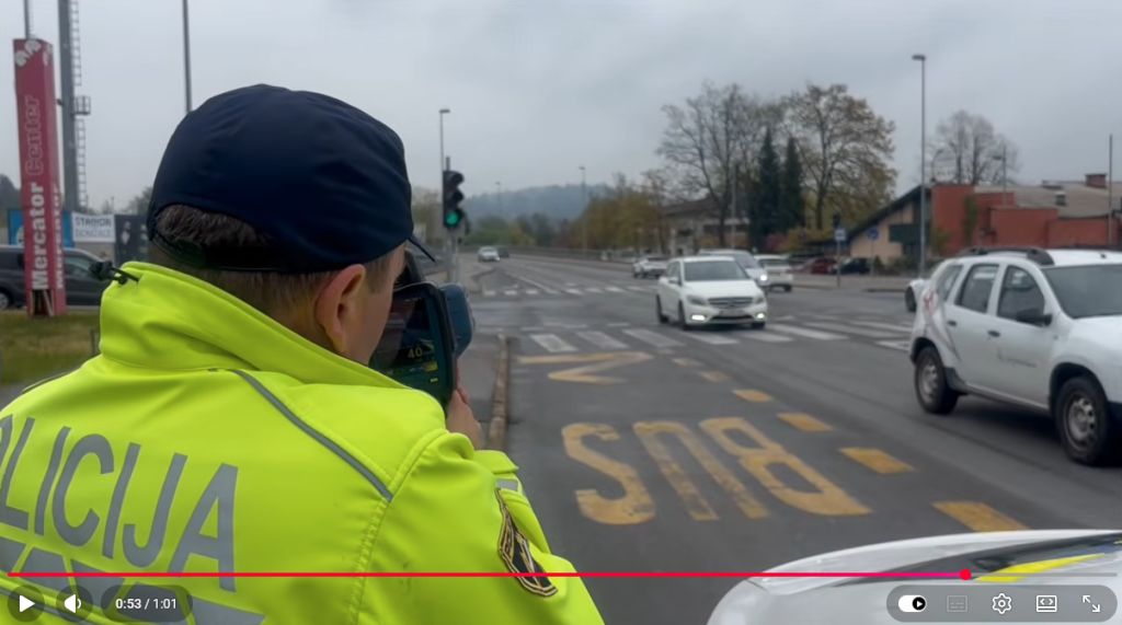 Izsek iz videa. V kadru je policist, ki gleda skozi ročni radar proti vozilom na cesti.